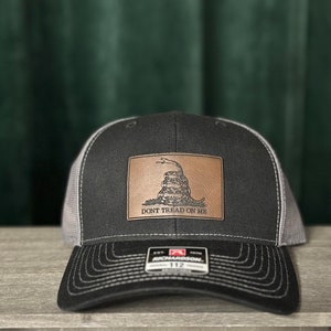 May include: A black and gray trucker hat with a brown leather patch featuring a coiled snake and the text "Don't Tread On Me".