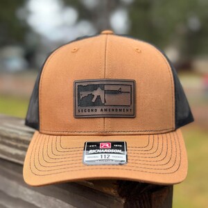 May include: A brown and black trucker hat with a brown leather patch that says "Second Amendment" and a silhouette of a rifle. The hat has a black mesh back and a brown adjustable strap with a black label that says "Richardson 112".