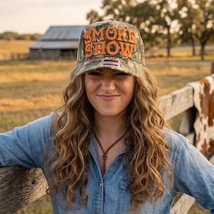May include: A camouflage baseball cap featuring the text SMOKE SHOW in raised orange lettering on the front panel. The cap has a structured crown, a curved brim, and a small rectangular label with red and white details positioned below the text.