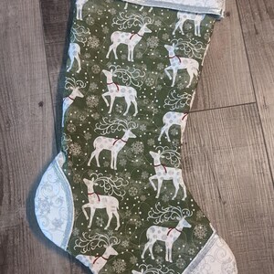 Könnte beinhalten: Ein grüner Weihnachtssocken mit einem weißen Rentiermuster und silbernem Besatz. Der Socken hat einen roten Knopf mit einem Schneeflockenmuster.