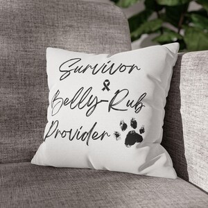 Op de afbeelding: Wit vierkant kussen met zwarte tekst die luidt "Survivor & Belly Rub Provider" en een pootafdruk. Het kussen ligt op een grijs gestoffeerde stoel.