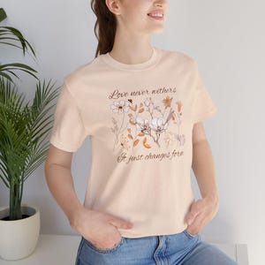 May include: A light peach-colored t-shirt with the text "Love never withers, It just changes form" and a floral design. The design features white, brown, and orange flowers. The shirt has short sleeves and a crew neck.
