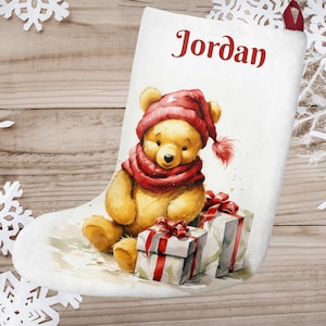 Può includere: Calza di Natale bianca con un'illustrazione di Winnie the Pooh. La calza presenta un orso giallo con un cappello e una sciarpa rossi, che tiene una scatola regalo. La calza è personalizzata con il nome "Jordan".