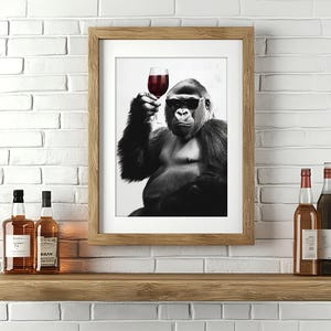May include: A black and white photograph of a gorilla holding a glass of red wine. The gorilla is sitting in a relaxed pose and looking directly at the viewer. The photograph is framed in a wooden frame.