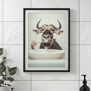 Puede incluir: Impresión artística enmarcada que presenta una imagen caprichosa de un ñu cepillándose los dientes sobre un lavabo blanco. El ñu tiene cuernos grandes y está situado sobre un fondo de azulejos. La obra de arte está enmarcada en un marco negro.