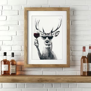May include: A framed black and white illustration of a deer wearing sunglasses and holding a glass of wine. The deer is looking to the right. The frame is brown wood.