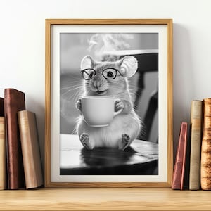 Puede incluir: Impresión en blanco y negro de un ratón con gafas sosteniendo una taza de café blanca. El ratón está sentado en una mesa y sale vapor de la taza. La impresión está enmarcada y se muestra en una estantería de madera con libros.