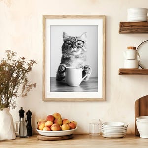 May include: Black and white photo of a cat wearing glasses and holding a white mug. The cat is sitting on a table and the photo is framed in a black frame.
