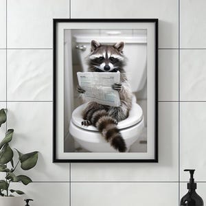 Puede incluir: Impresión enmarcada de un mapache sentado en un inodoro, leyendo un periódico. El mapache tiene una máscara negra distintiva y una cola tupida. La impresión está en un marco negro y cuelga de una pared de azulejos blancos.