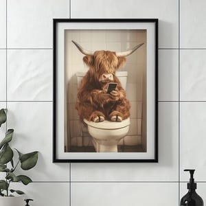 May include: A framed print of a Highland cow sitting on a toilet and using a smartphone. The cow is brown and white with long hair. The toilet is white and the background is a white tiled wall.