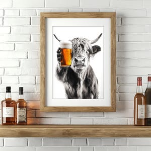 Pode incluir: Uma fotografia a preto e branco de uma vaca das terras altas segurando um copo de cerveja. A vaca está a olhar diretamente para a câmara. A fotografia está enquadrada numa moldura de madeira castanho.