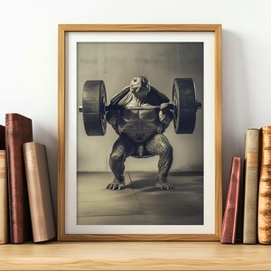Op de afbeelding: Een ingelijste foto van een schildpad die een halterstang optilt. De schildpad staat in een squat-positie met gebogen benen en zijn schild naar de camera gericht. De halterstang is van metaal en heeft aan beide uiteinden gewichtsplaten. De achtergrond is lichtbruin.