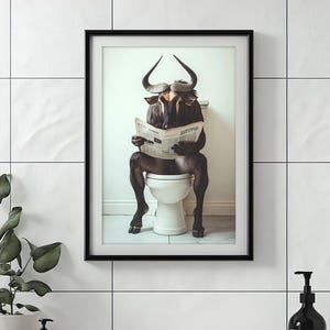Puede incluir: Impresión artística enmarcada que presenta una imagen caprichosa de un ñu leyendo un periódico mientras está sentado en un inodoro. La obra de arte tiene un marco negro y se muestra contra una pared de azulejos blancos. El ñu está representado de forma realista.