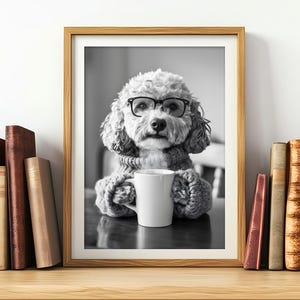 May include: A black and white photo of a dog wearing glasses and a sweater, holding a white mug. The dog is sitting at a table with a book in the background.