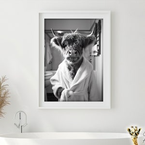 May include: Black and white photograph of a Highland cow wearing a white bathrobe. The cow is standing in a bathroom with a towel rack and a toilet visible in the background.