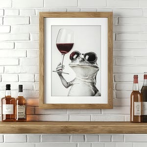 Può includere: Una stampa incorniciata di una rana con occhiali da sole che tiene un bicchiere di vino rosso. La stampa è appesa a un muro di mattoni bianchi sopra uno scaffale di legno con due bottiglie di liquore.