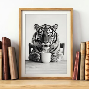 May include: A black and white photograph of a tiger holding a white mug. The tiger is seated at a table with a white background. The tiger is looking directly at the camera.