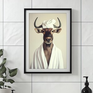 May include: Framed art print featuring a wildebeest wearing a white robe and a chef's hat. The animal has large horns and is centered against a light background. The artwork is in a black frame and is displayed on a white tiled wall.