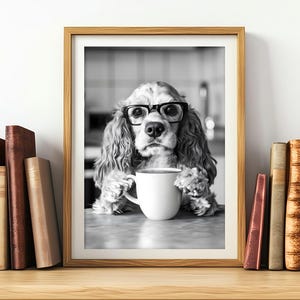 Puede incluir: Una fotografía en blanco y negro de un perro con gafas y sosteniendo una taza de café blanca. El perro está sentado en una mesa con un libro en el fondo.