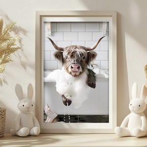 May include: A framed print depicting a Highland cow in a bubble bath. The cow's head and shoulders are visible, with its long, shaggy fur and large horns. The background shows a white tiled wall. Decorative items, including a bunny figurine and a basket, are in the foreground.