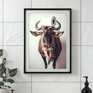 Puede incluir: Impresión artística enmarcada que presenta un ñu con un rollo de papel higiénico equilibrado en sus cuernos. La obra de arte está en un marco negro y tiene un fondo blanco. El ñu es marrón y canela con cuernos grandes.