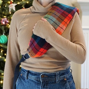 May include: A colorful plaid shoulder wrap in shades of red, orange, blue, and green. The wrap has a black strap with a metal ring. The background includes a Christmas tree with ornaments.
