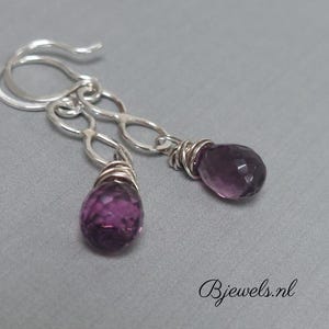 Op de afbeelding: Een paar zilveren oorbellen met paarse edelsteen druppels. De oorbellen hebben een delicaat kettingontwerp en zijn versierd met zilverdraad.