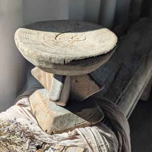 Puede incluir: Un pedestal rústico de madera con una parte superior redonda y una base geométrica. El pedestal está hecho de madera envejecida, mostrando la veta y textura natural. La pieza se exhibe sobre una superficie de madera con tela drapeada, creando una estética natural y terrosa.
