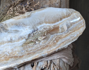 May include: A large, oblong onyx tray with cream and brown veining. Dried flowers and grasses are arranged on the tray. The tray rests on a weathered, rustic wooden surface, with a neutral-colored fabric draped beneath.