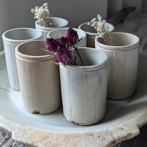 Pode incluir: Um conjunto de seis potes cilíndricos antigos de grés, em tons de creme e branco sujo, dispostos numa bandeja com efeito mármore. Alguns potes contêm flores secas e rosas roxas escuras. Os potes apresentam detalhes de nervuras verticais.