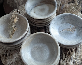 May include: A collection of small, round, light grey stone bowls with natural veining and smooth, matte surfaces. The bowls are stacked in three separate piles, with dried botanical sprigs placed inside two of the bowls as decorative accents.