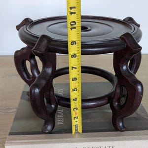 Puede incluir: Un soporte de exhibici&oacute;n de madera marr&oacute;n oscuro, de aproximadamente 15 cm de altura, con una parte superior circular y patas curvas ornamentadas. El soporte est&aacute; dise&ntilde;ado para sostener un objeto decorativo. El soporte est&aacute; sobre un libro con las palabras "RURAL RETREATS" en la portada.