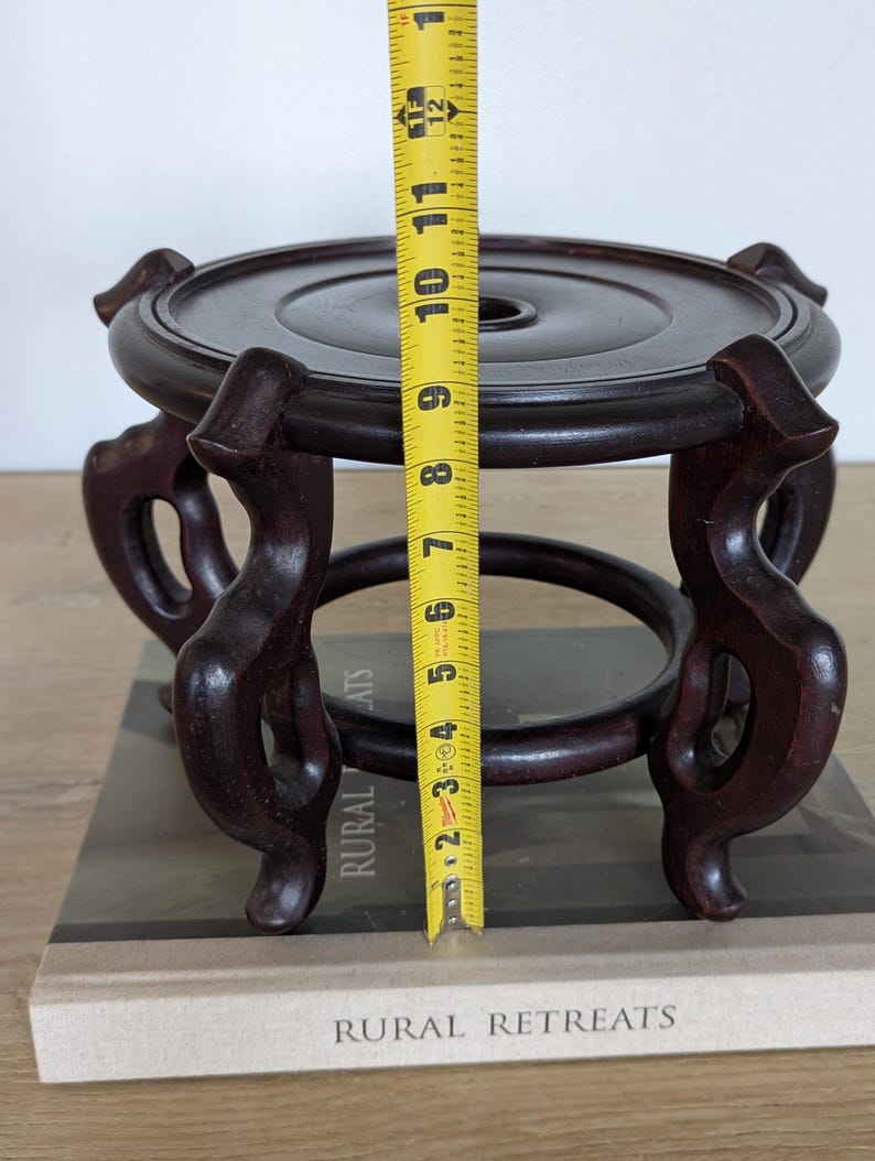 Puede incluir: Un soporte de exhibici&oacute;n de madera marr&oacute;n oscuro, de aproximadamente 15 cm de altura, con una parte superior circular y patas curvas ornamentadas. El soporte est&aacute; dise&ntilde;ado para sostener un objeto decorativo. El soporte est&aacute; sobre un libro con las palabras "RURAL RETREATS" en la portada.