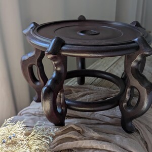 Puede incluir: Un soporte de exhibici&oacute;n de madera marr&oacute;n oscuro con una parte superior redonda y patas curvas ornamentadas. El soporte tiene una base circular y est&aacute; dise&ntilde;ado para sostener un objeto decorativo. El soporte tiene aproximadamente 15 cm de di&aacute;metro.