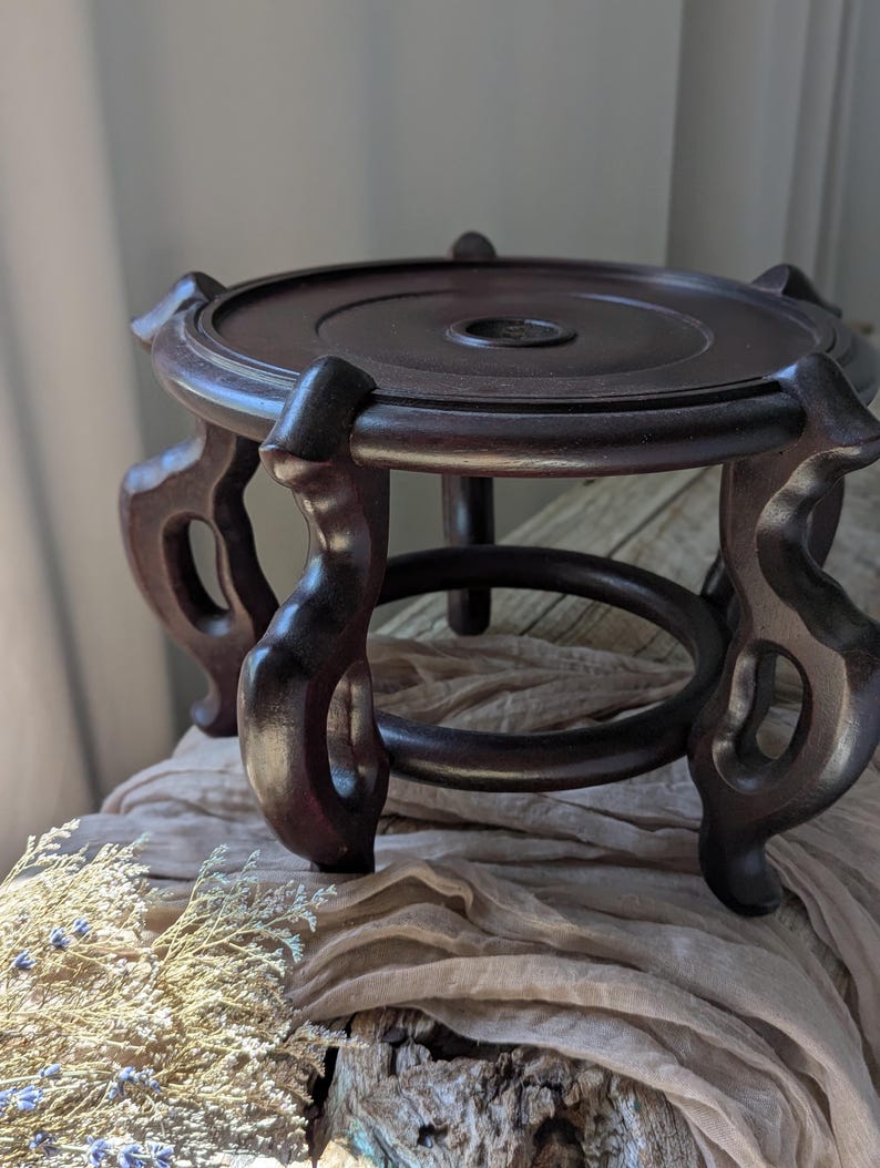 Puede incluir: Un soporte de exhibici&oacute;n de madera marr&oacute;n oscuro con una parte superior redonda y patas curvas ornamentadas. El soporte tiene una base circular y est&aacute; dise&ntilde;ado para sostener un objeto decorativo. El soporte tiene aproximadamente 15 cm de di&aacute;metro.