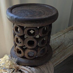 May include: A dark brown wooden stool with a circular top and base. The body of the stool features a series of carved-out circles. The stool is placed on a wooden surface with a draped fabric and dried flowers.