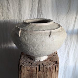 May include: A light gray, textured ceramic vase with a rounded body and a wide, slightly flared rim. The vase sits atop a weathered, rough-hewn wooden block. The background is a crumpled white fabric.