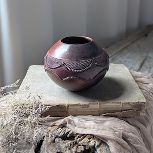 May include: A spherical dark brown clay vessel with a smooth finish and a wide circular opening. The midsection features a repeating embossed scalloped pattern with fine textured lines, creating a decorative band around the centre of the pot.