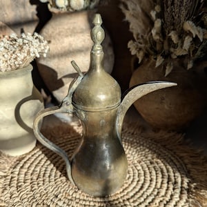 May include: Antique brass coffee pot with a long spout and a curved handle. The pot has a decorative finial on the lid and is set on a woven mat. The pot has a vintage look and feel.