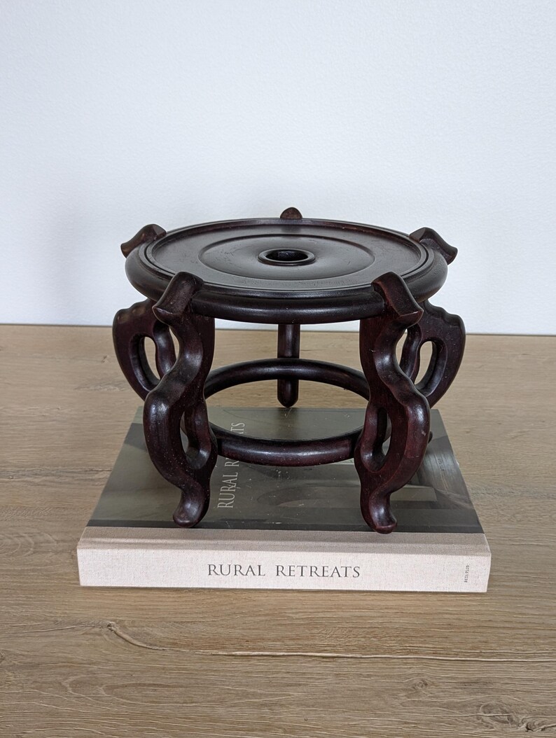 Puede incluir: Un soporte de exhibici&oacute;n de madera marr&oacute;n oscuro con una parte superior circular y patas curvas ornamentadas. El soporte mide aproximadamente 15 cm de di&aacute;metro y 10 cm de alto. Est&aacute; colocado sobre un libro titulado "Rural Retreats".