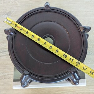 Puede incluir: Un soporte de madera marr&oacute;n oscuro con una parte superior circular y tres patas decorativas. El soporte tiene aproximadamente 33 cm de di&aacute;metro. Una cinta m&eacute;trica amarilla est&aacute; colocada sobre la parte superior del soporte.