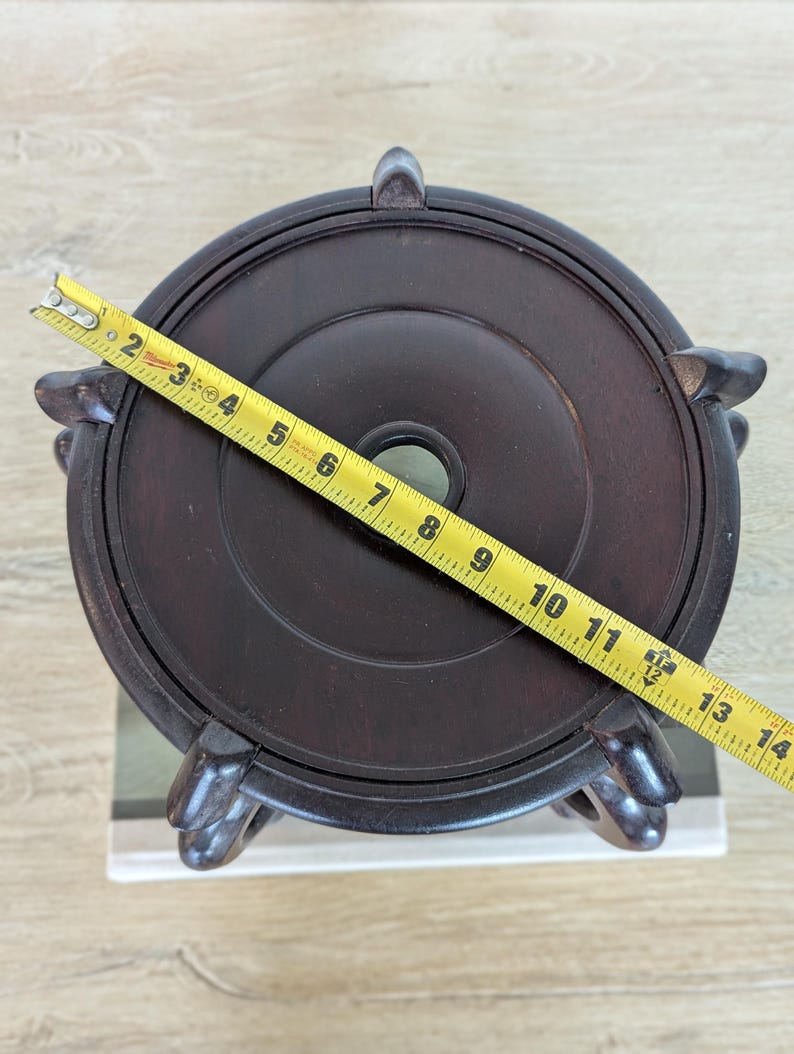 Puede incluir: Un soporte de madera marr&oacute;n oscuro con una parte superior circular y tres patas decorativas. El soporte tiene aproximadamente 33 cm de di&aacute;metro. Una cinta m&eacute;trica amarilla est&aacute; colocada sobre la parte superior del soporte.