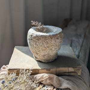 May include: A weathered, stone-like vessel with a wide opening, possibly a mortar or small planter, sits atop an aged book. Dried sprigs of plant life are inside the vessel. The scene is set on a rustic wooden surface with a neutral-toned fabric.