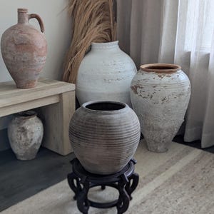 Puede incluir: Una colecci&oacute;n de jarrones r&uacute;sticos y texturizados de varias formas y tama&ntilde;os. La paleta de colores incluye tonos de blanco, marr&oacute;n y gris. Un jarr&oacute;n tiene un asa, otro se apoya en un soporte de madera oscura. La escena est&aacute; sobre una alfombra neutra.