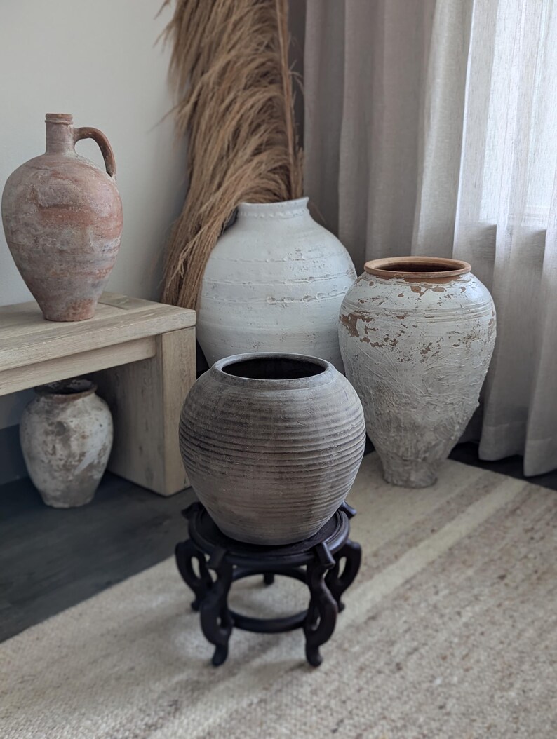 Puede incluir: Una colecci&oacute;n de jarrones r&uacute;sticos y texturizados de varias formas y tama&ntilde;os. La paleta de colores incluye tonos de blanco, marr&oacute;n y gris. Un jarr&oacute;n tiene un asa, otro se apoya en un soporte de madera oscura. La escena est&aacute; sobre una alfombra neutra.