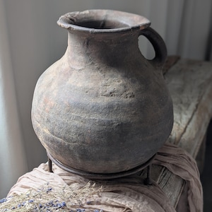 May include: A rustic, dark brown earthenware jug with a rounded body and a handle. The jug sits on a metal stand, and is displayed on a weathered wooden surface with a draped fabric and dried flowers.