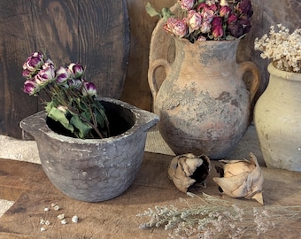 May include: A rustic still life composition. It features a wooden mortar with dried roses, a weathered terracotta vase with roses, and a smaller vase. The arrangement is on a wooden plank, with dried floral accents.