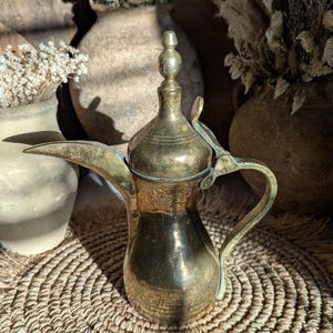 May include: A vintage brass coffee pot with a long spout and ornate handle. The pot has a decorative lid and sits on a woven mat. Dried flowers in ceramic vases are in the background, creating a warm, rustic aesthetic.
