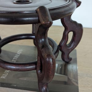 Puede incluir: Un soporte de madera oscura y ornamentada, con una parte superior circular y patas decorativas y curvas. El soporte tiene un acabado rico y pulido y est&aacute; dise&ntilde;ado para sostener un objeto peque&ntilde;o. Con un agujero central.
