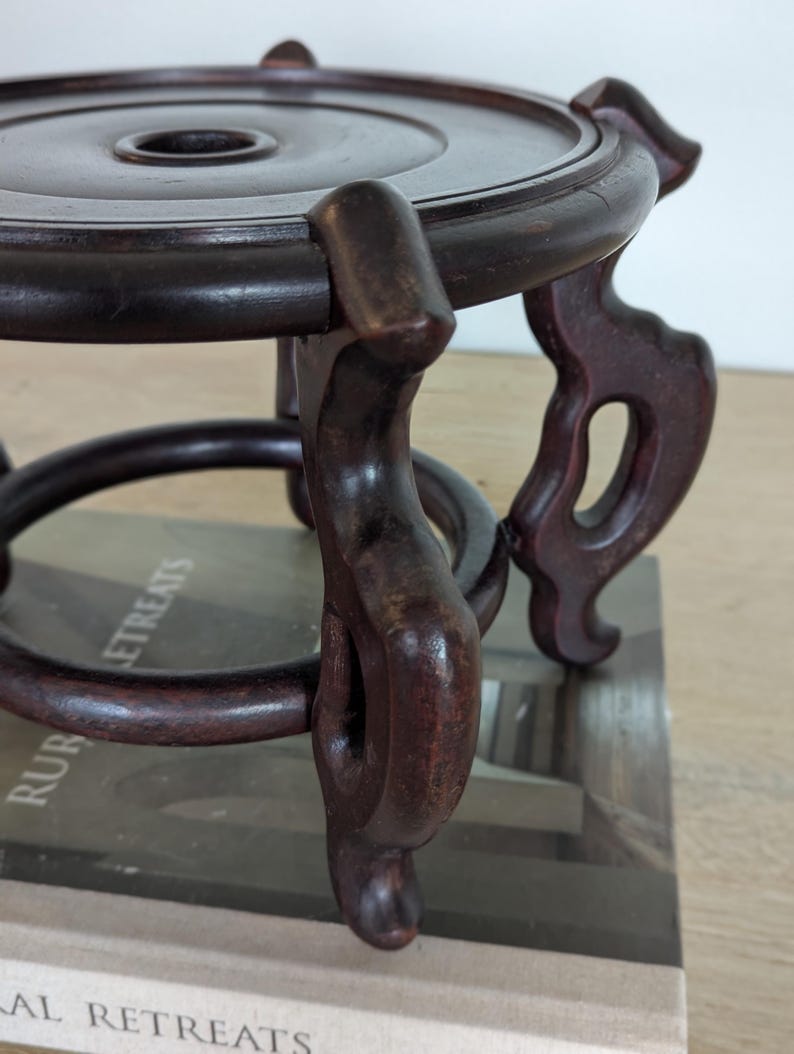 Puede incluir: Un soporte de madera oscura y ornamentada, con una parte superior circular y patas decorativas y curvas. El soporte tiene un acabado rico y pulido y est&aacute; dise&ntilde;ado para sostener un objeto peque&ntilde;o. Con un agujero central.